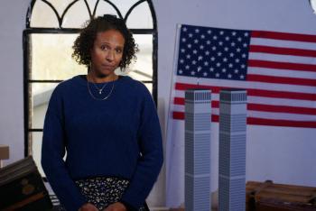 Presentatrice Astrid staat voor een raam. Rechts van haar staan de Twin Towers en erboven een Amerikaanse vlag.